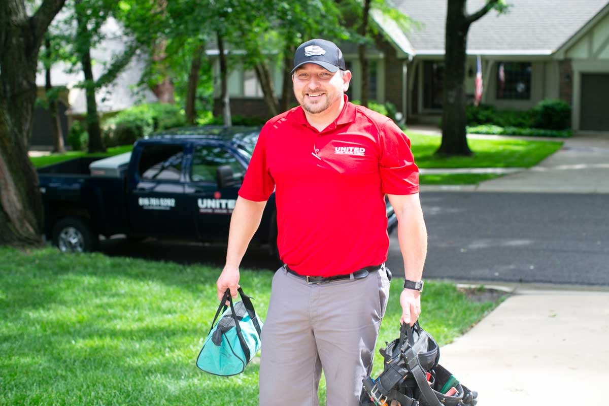 United Heating technician wearing a bright red collared uniform shirt, carrying tools, and smiling as he walks up the driveway toward a client's home